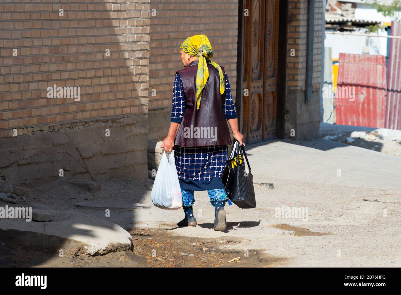 Die alte asiatin, die von hinten gesehen wurde, hält Taschen Handtasche und trägt traditionelle Kleidung mit Stempel und farbenfrohem Schal in Samarkand, Usbekistan. Stockfoto