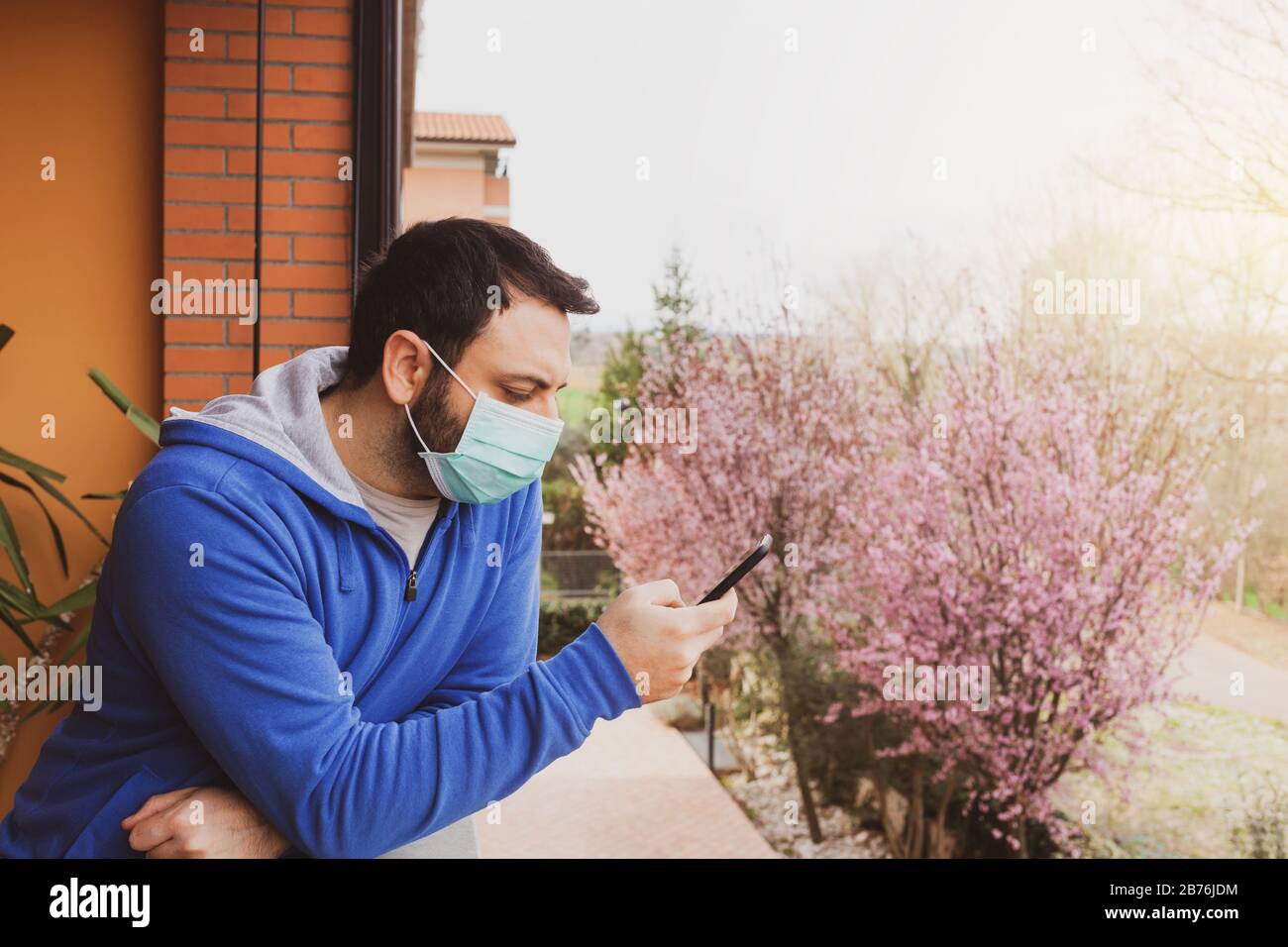Junger kaukasischer Mann mit Maske, der während der Quarantäne mit dem Handy auf die Heimatterrasse blickt, wegen Coronavirus Covid19-Pandemie. Stockfoto