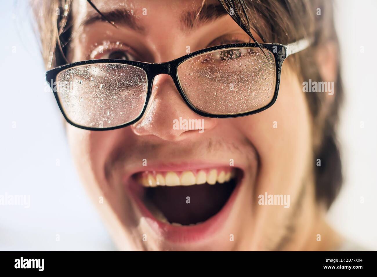 Ein sehr überraschter junger Mann in einer Brille, dessen Glas mit kleinen Wassertropfen bedeckt ist und von hellen Sonnenstrahlen beleuchtet wird. Spiele mit Stockfoto