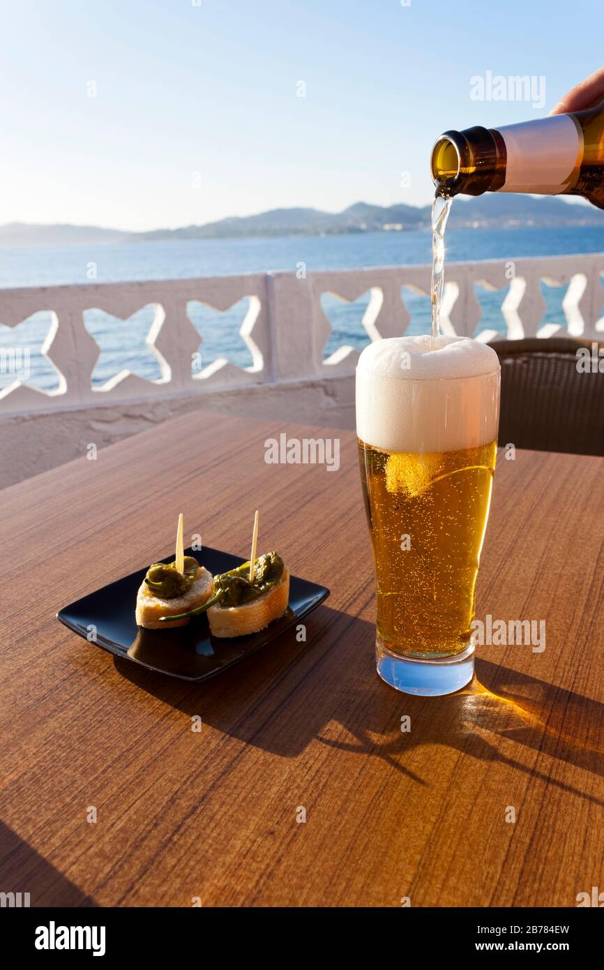 Gießen kaltes Bier aus der Flasche in ein Glas und einen Teller mit grünem Paprika in der Tabelle eine Terrasse direkt am Meer in Spanien. Stockfoto