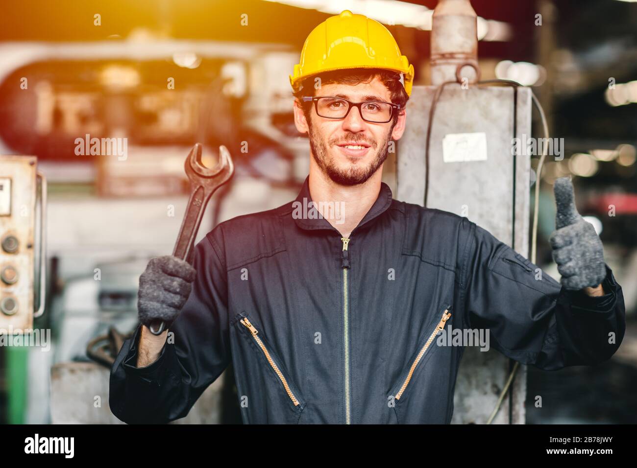 Portrait junger amerikanischer glücklicher Arbeiter freut sich, lächelnd in einer schweren Industriefabrik zu arbeiten.Daumen hoch mit großem Schraubenschlüssel. Stockfoto