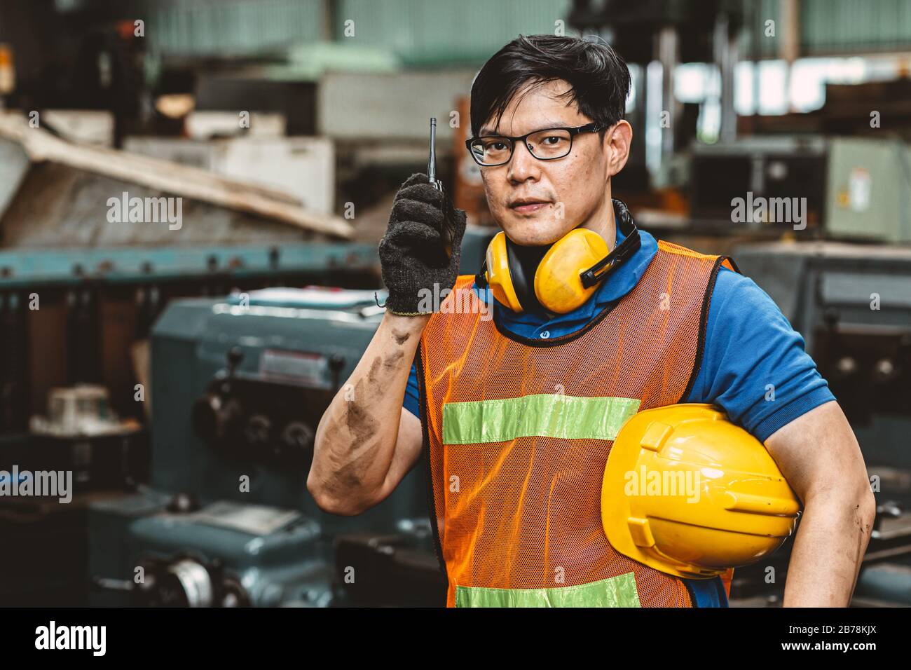 Asiatischer Techniker mit Schutzanzug arbeitet im Werk hart an der Kommunikation mit dem Radio, das Kamera und Lächeln sieht. Stockfoto