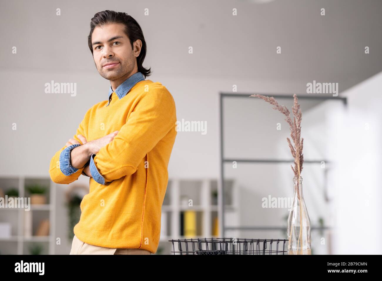 Junger, eleganter Geschäftsmann in gelbem Pullover und Denimhemd, der Sie im Büro am Arbeitsplatz betrachtet Stockfoto