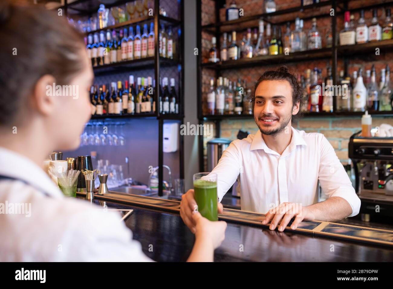 Fröhlicher junger Barmann, der ein Glas Gemüsecocktail oder Smoothie für den Kunden hält, während er es über die Theke an seinen Kollegen weitergibt Stockfoto