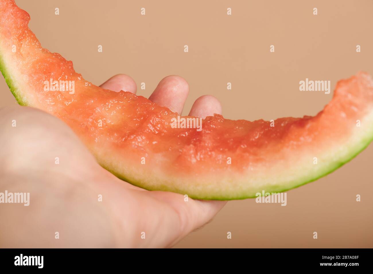 Halten Sie die verzehrte Wassermelonenscheibe in Nahaufnahme auf beigefarbenem Farbhintergrund Stockfoto