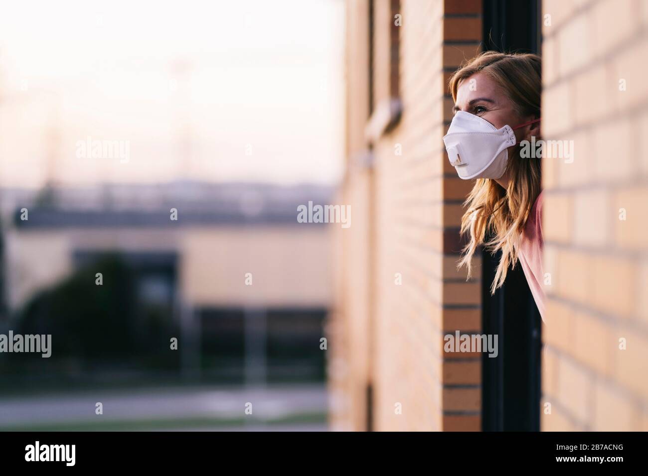 Stockfoto der Frau, die aus dem Hausfenster schaut. Sie trägt eine Schutzmaske zur Vorbeugung gegen einen Virus. Coronavirus Konzept Stockfoto
