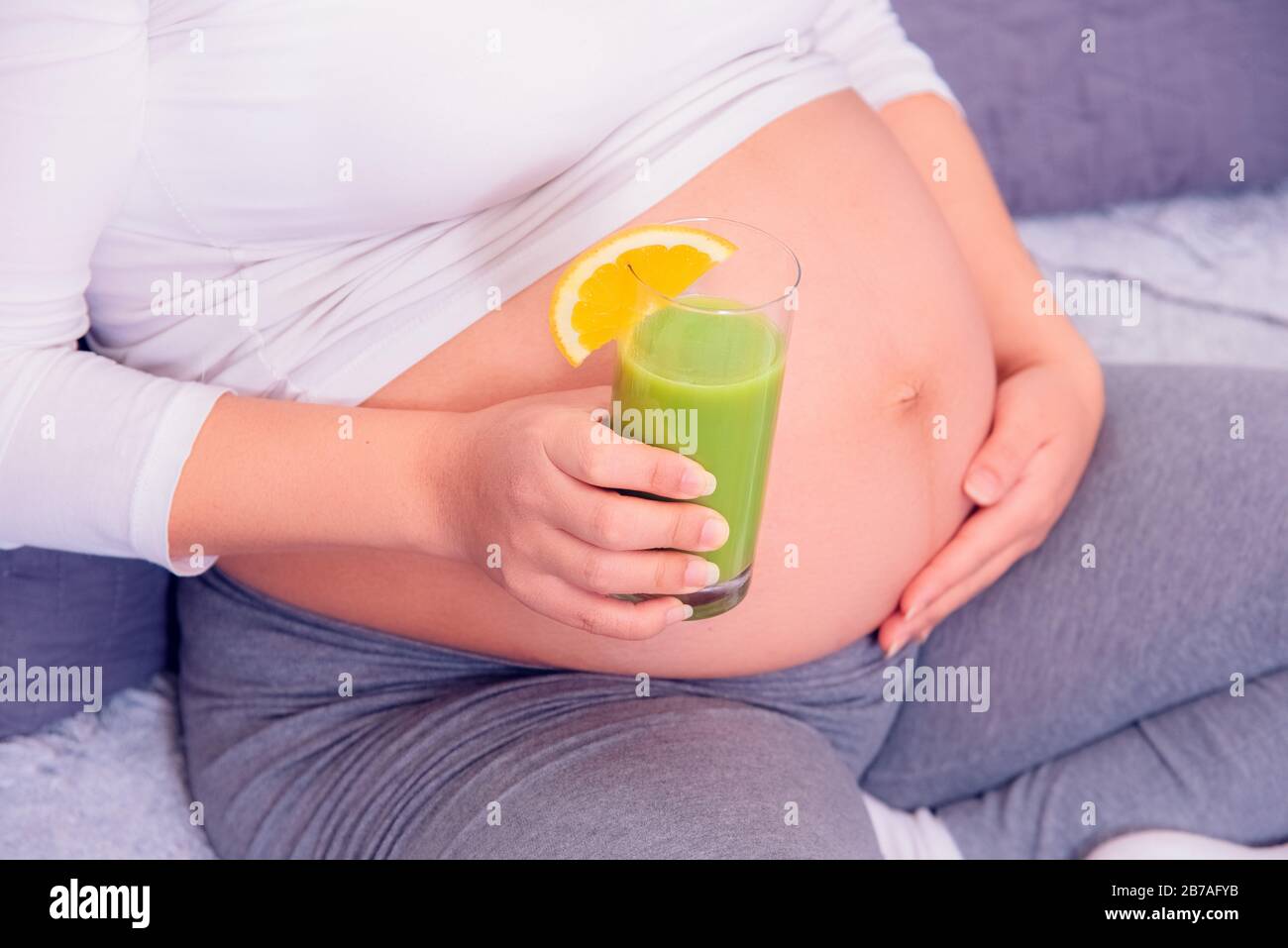 Schwangere Frau, die ihren gesunden grünen Biosaft genießt und sich zu Hause auf der Couch entspannt. Stockfoto