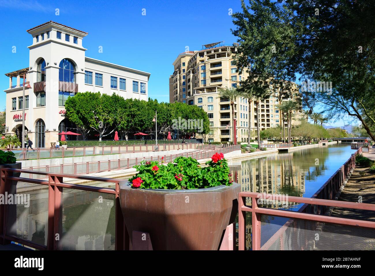 Gehobene Eigentumswohnungen, die in der Uferpromenade entlang des AZ-Kanals mit Restaurants und Red Begonias in Betonpflanzern in Scottsdale, AZ, reflektieren Stockfoto