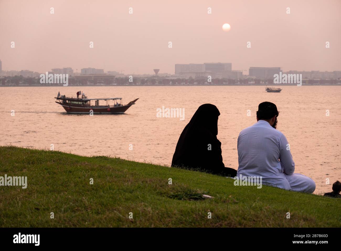 Ein muslimischer Mann und eine Frau in einer Burka und Taube beobachten den Sonnenuntergang, während ein Dhow-Boot über den Hafen in Doha, Katar, segelt Stockfoto