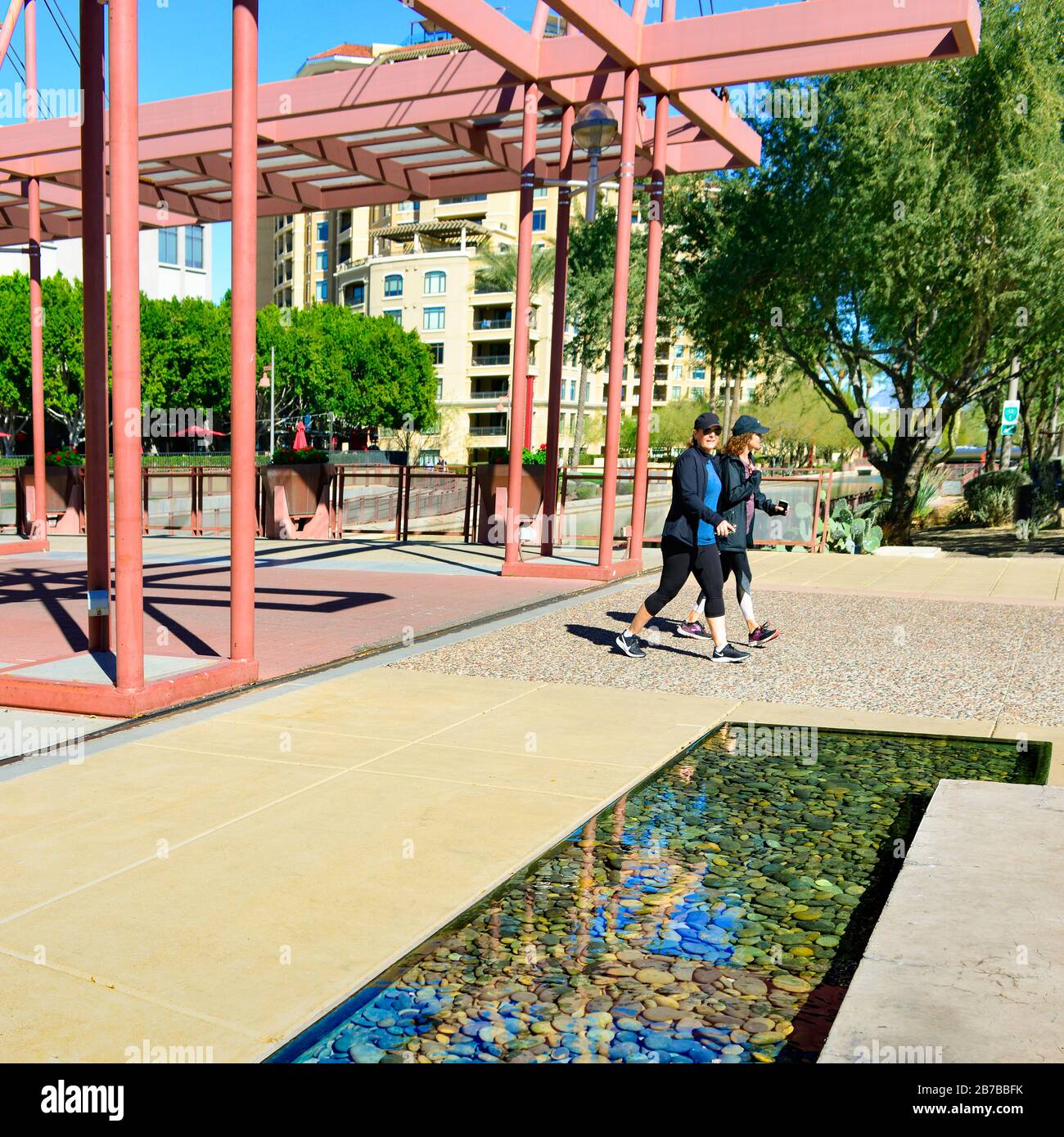 Zwei Frauen in sportlicher Kleidung und Turnschuhe gehen entlang des AZ-Kanalbereichs an der Scottsdale Waterfront mit modernen industriellen Akzenten und Condos Stockfoto