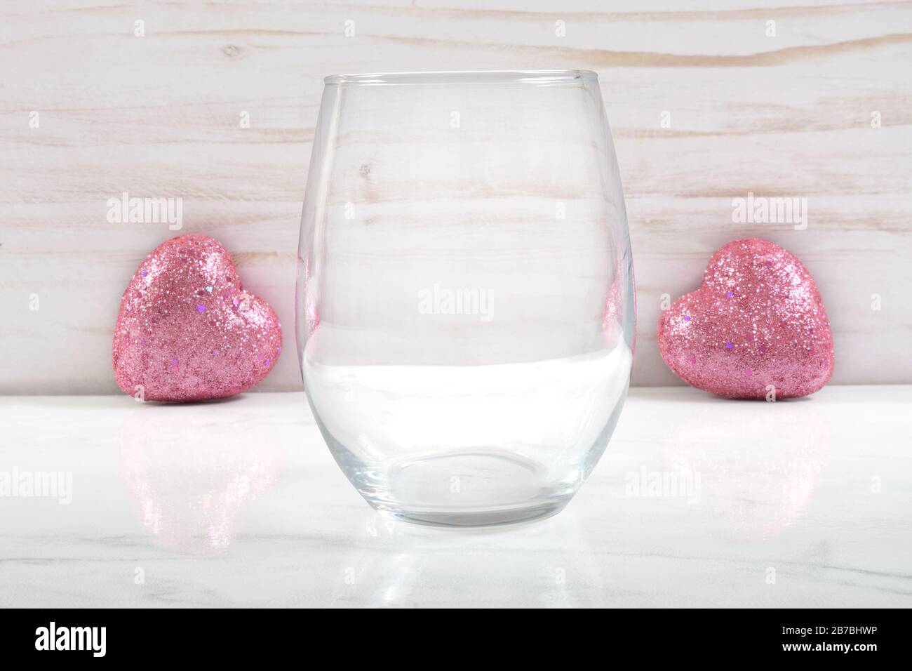 Ein wunderschönes, stielloses Weinglas ruht romantisch auf einem weißen Marmorhintergrund. Zwei rosarote Herzen funkeln im Hintergrund. Stockfoto