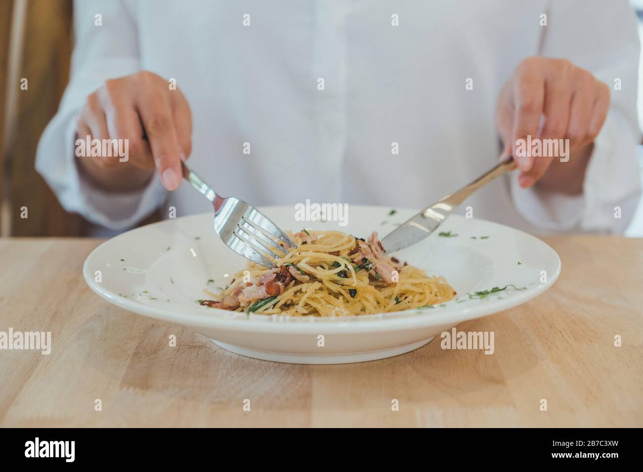 Nahaufnahme einer Frau, die mit Messer und Gabel würzige Speckspaghetti auf Holztisch isst Stockfoto