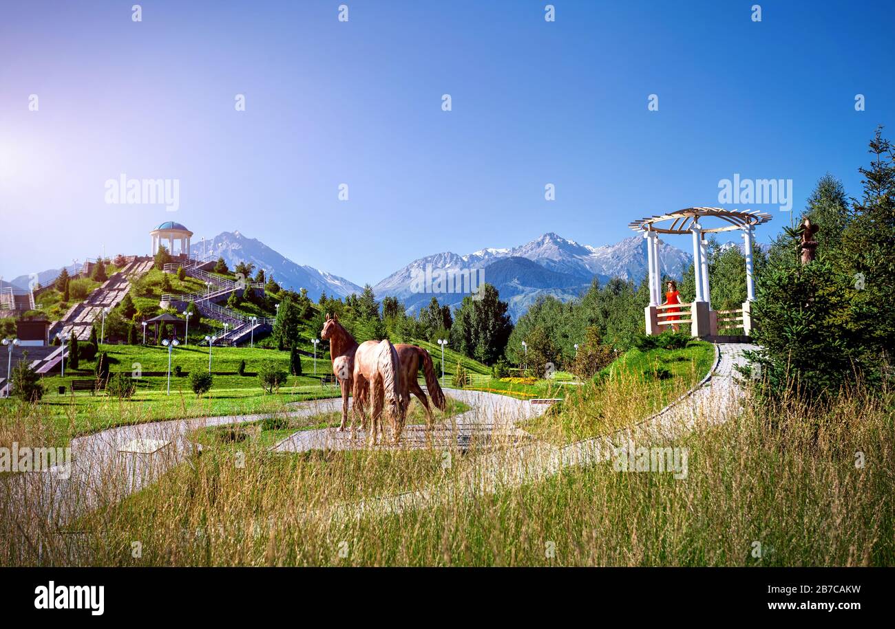 Frau in orangefarbenem Kleid im Park mit Pferdestatue und Bergen in Almaty, Kasachstan Stockfoto