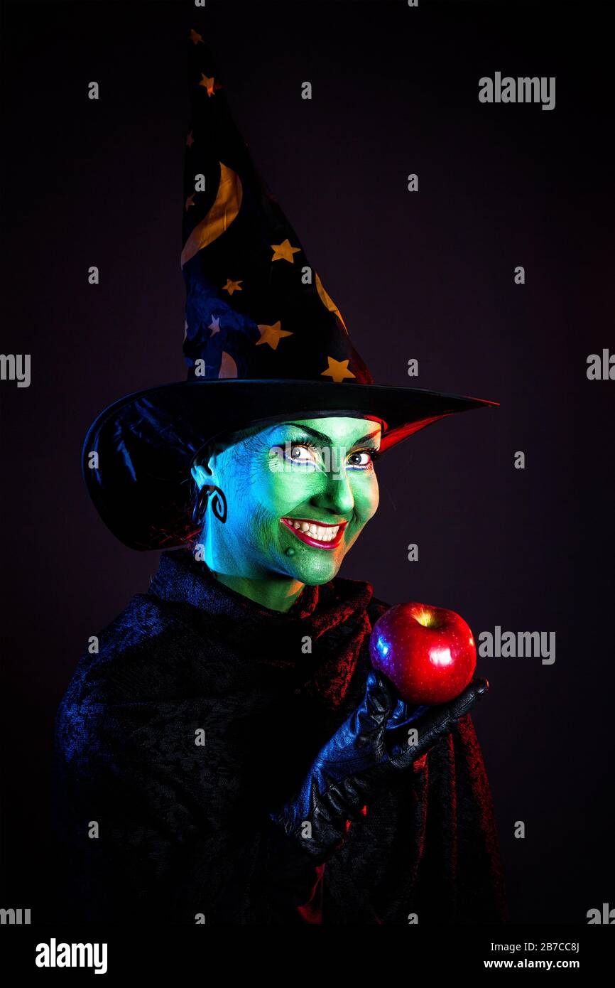 Hexe mit grüner Haut, die roten apfel hält und an Halloween auf dunklem Hintergrund lächelt Stockfoto