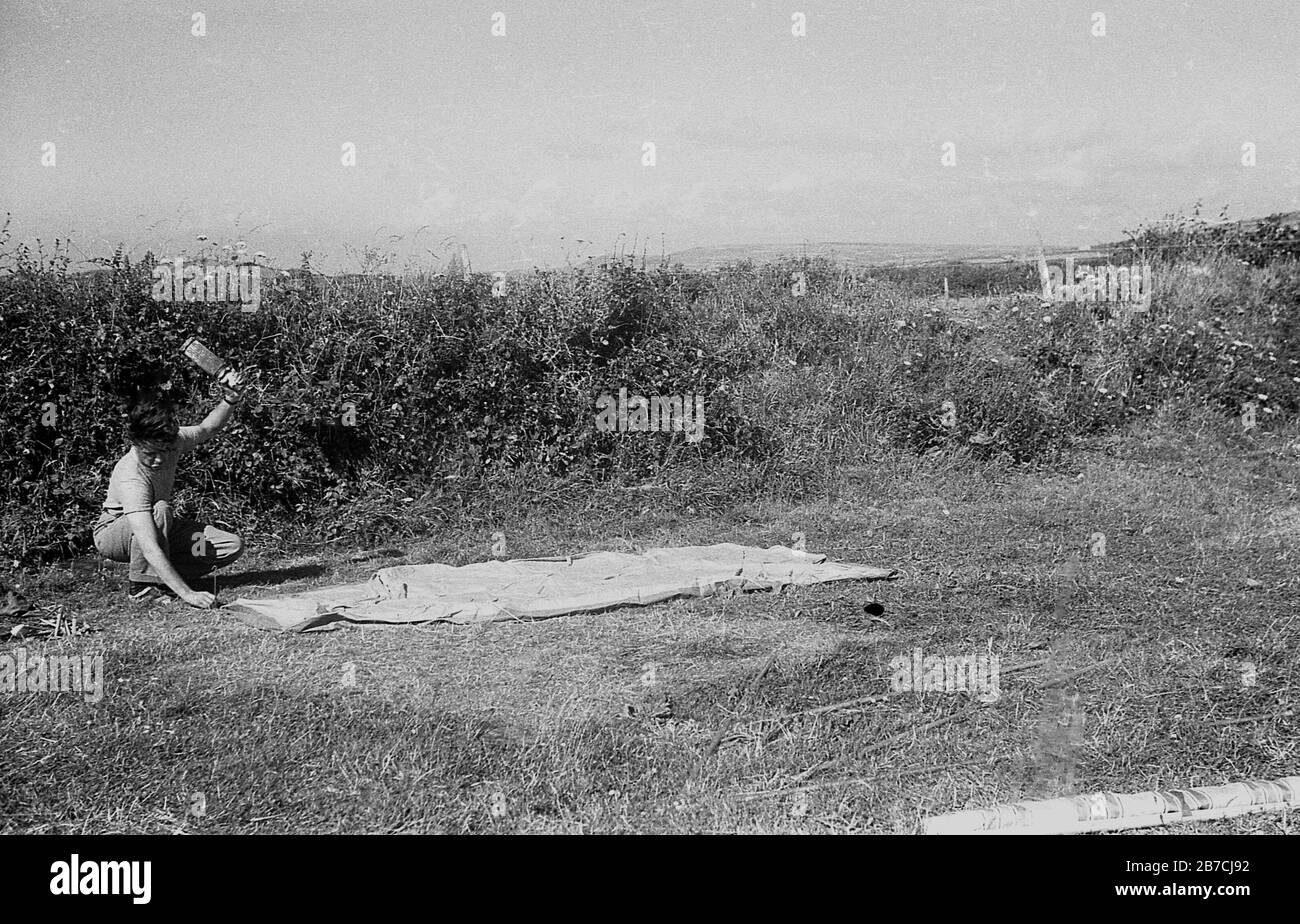 1950er Jahre, historisch, an der Küste, ein männliches Wohnmobil am Rand eines mit Hecke umringten Feldes, das einen Bodenblech eines Zeltes mit einem Holzhammer auf das Gras fixiert, um in die Pflöcke, Cornwall, England, Großbritannien zu schlagen. Stockfoto