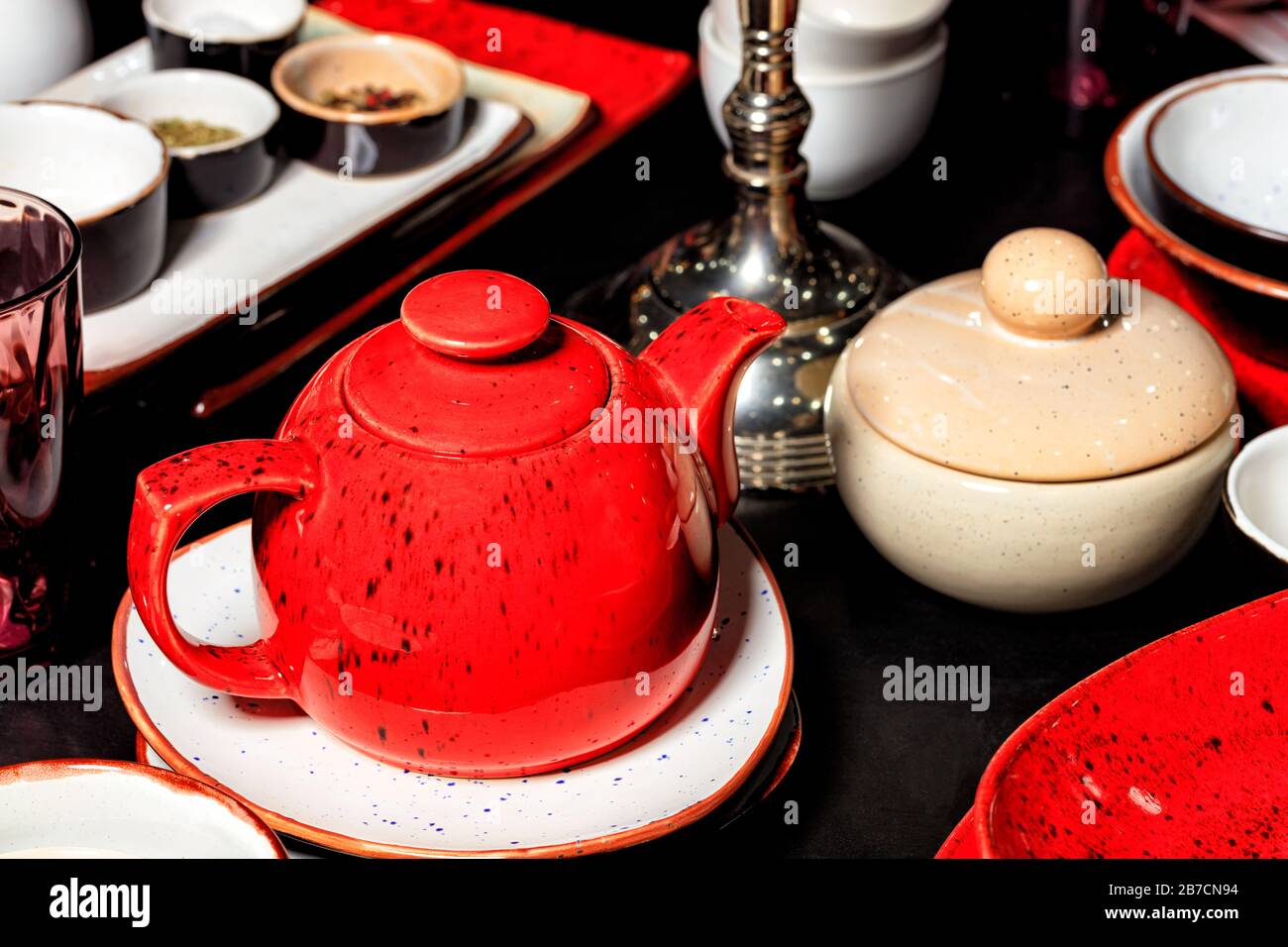 Glänzende Teekanne aus rotem Porzellan mit schwarzen Punkten auf einem schwarzen Tisch, unter anderem mit Teeutensilien, Tellern und handgefertigten Behältern. Selektiver Fokus. Stockfoto