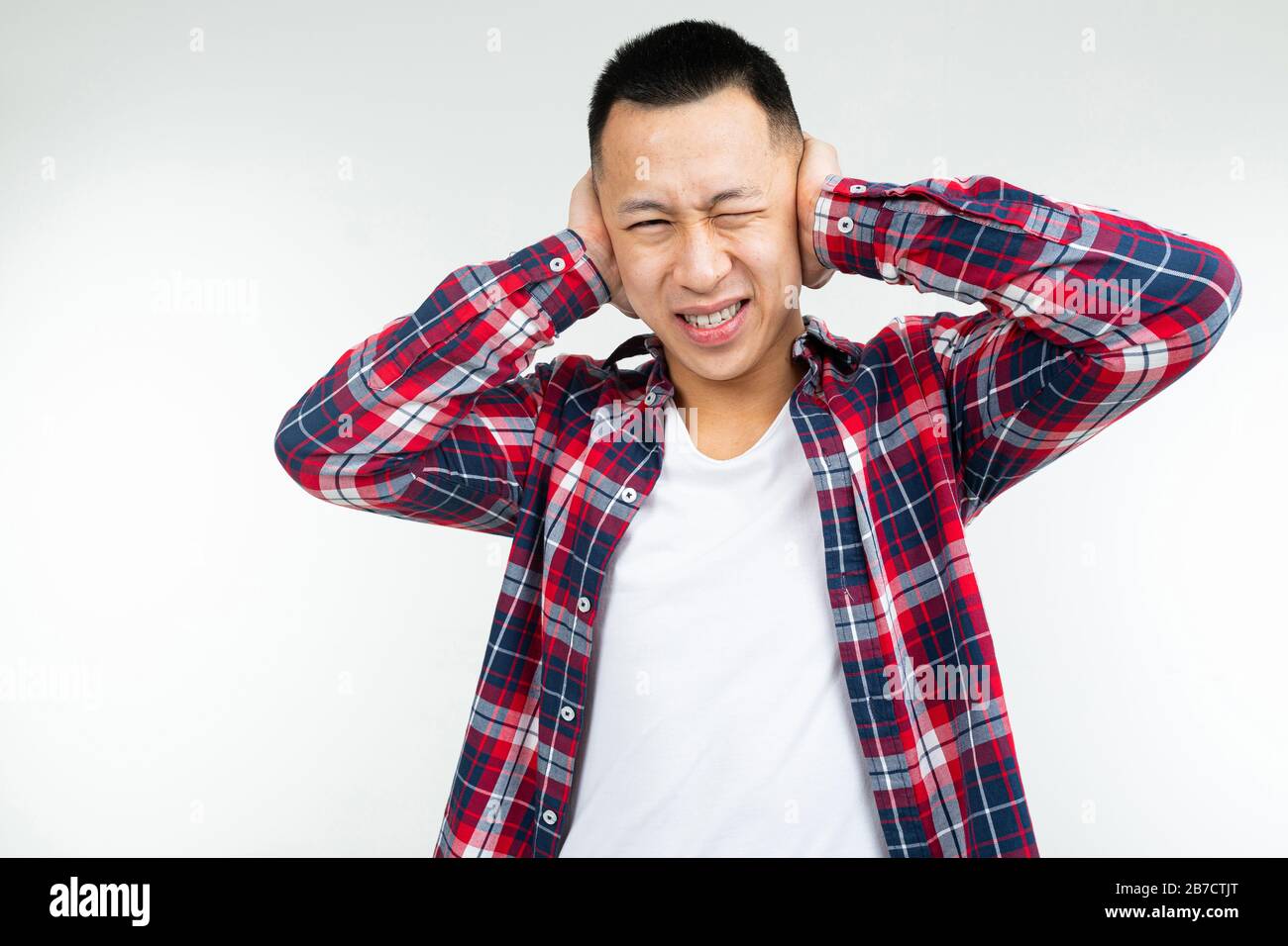 Gutaussehender asiate in einem karierten Hemd, das weit offen ist, bedeckt seine Ohren vor Lärm auf einem weißen Studiohintergrund Stockfoto