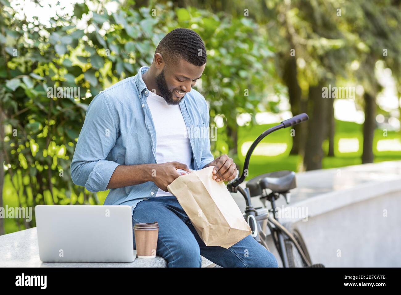 Mittagessen Im Park. Black Guy, Der Im Freien Mit Laptop Isst Und Ausruht Stockfoto