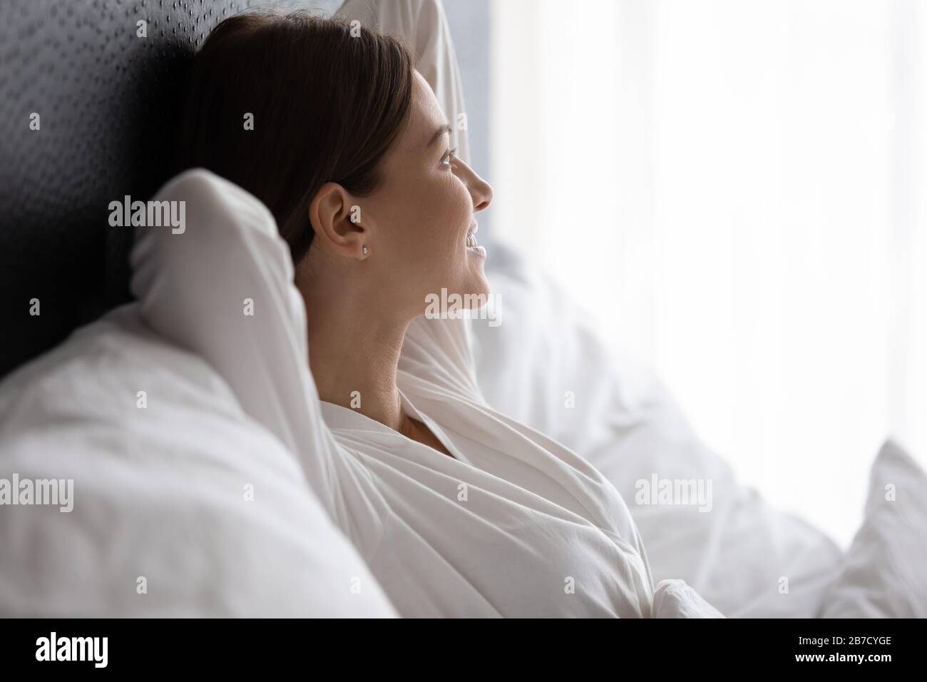 Lächelnde, attraktive junge Brunette-Frau, die sich am Morgen im Bett genießt. Stockfoto