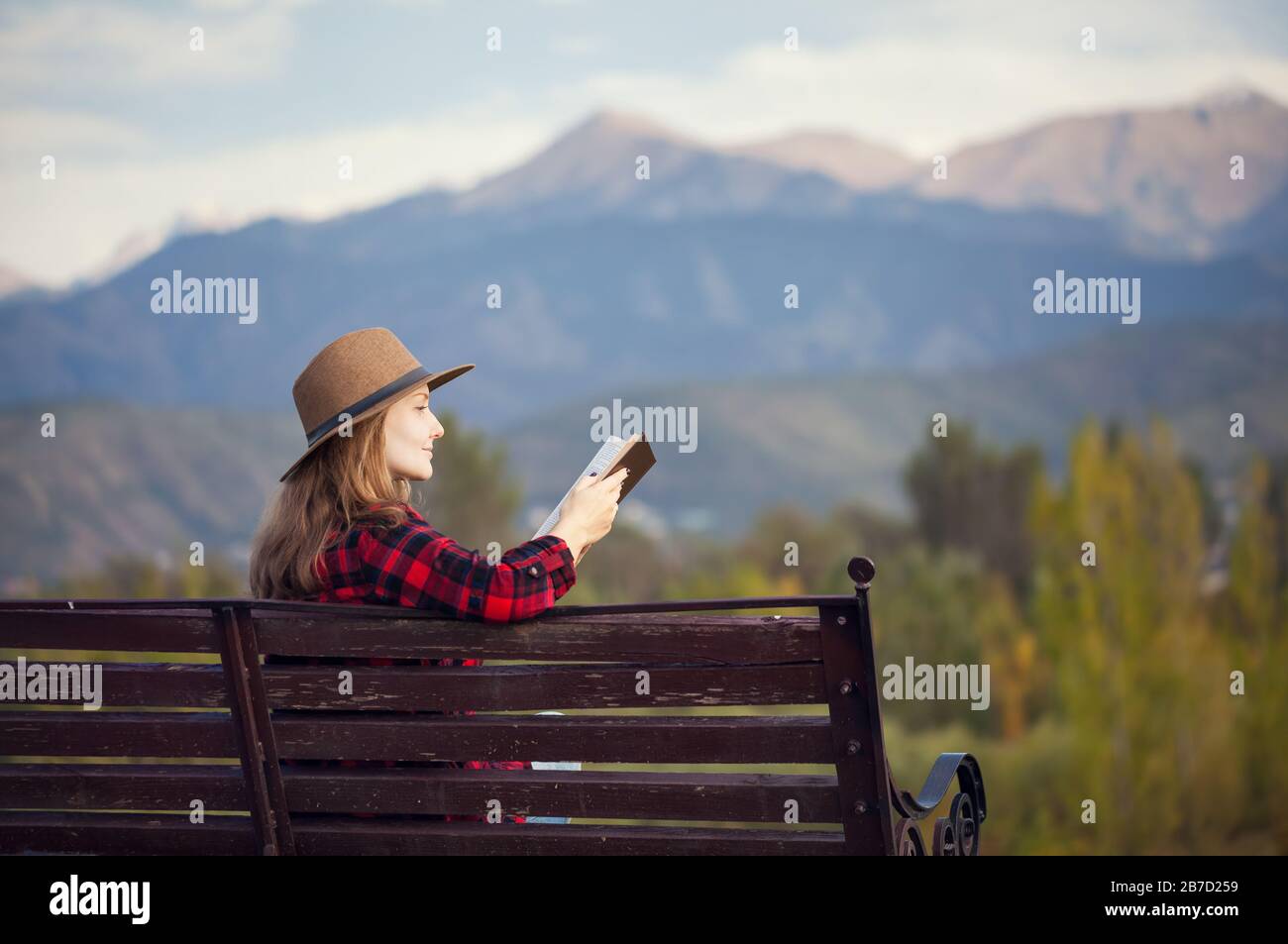 Frau in Rot kariertes Hemd und Hut sitzt auf der Holzbank und Lesebuch im herbstlichen Wald und Berge-Hintergrund Stockfoto