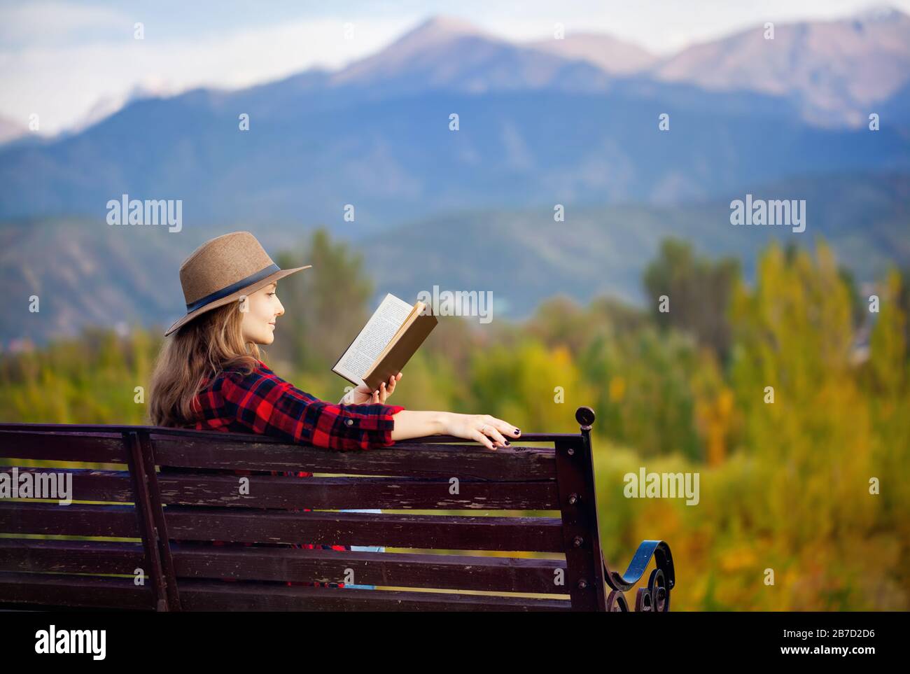 Frau in Rot kariertes Hemd und Hut sitzt auf der Holzbank und Lesebuch im herbstlichen Wald und Berge-Hintergrund Stockfoto