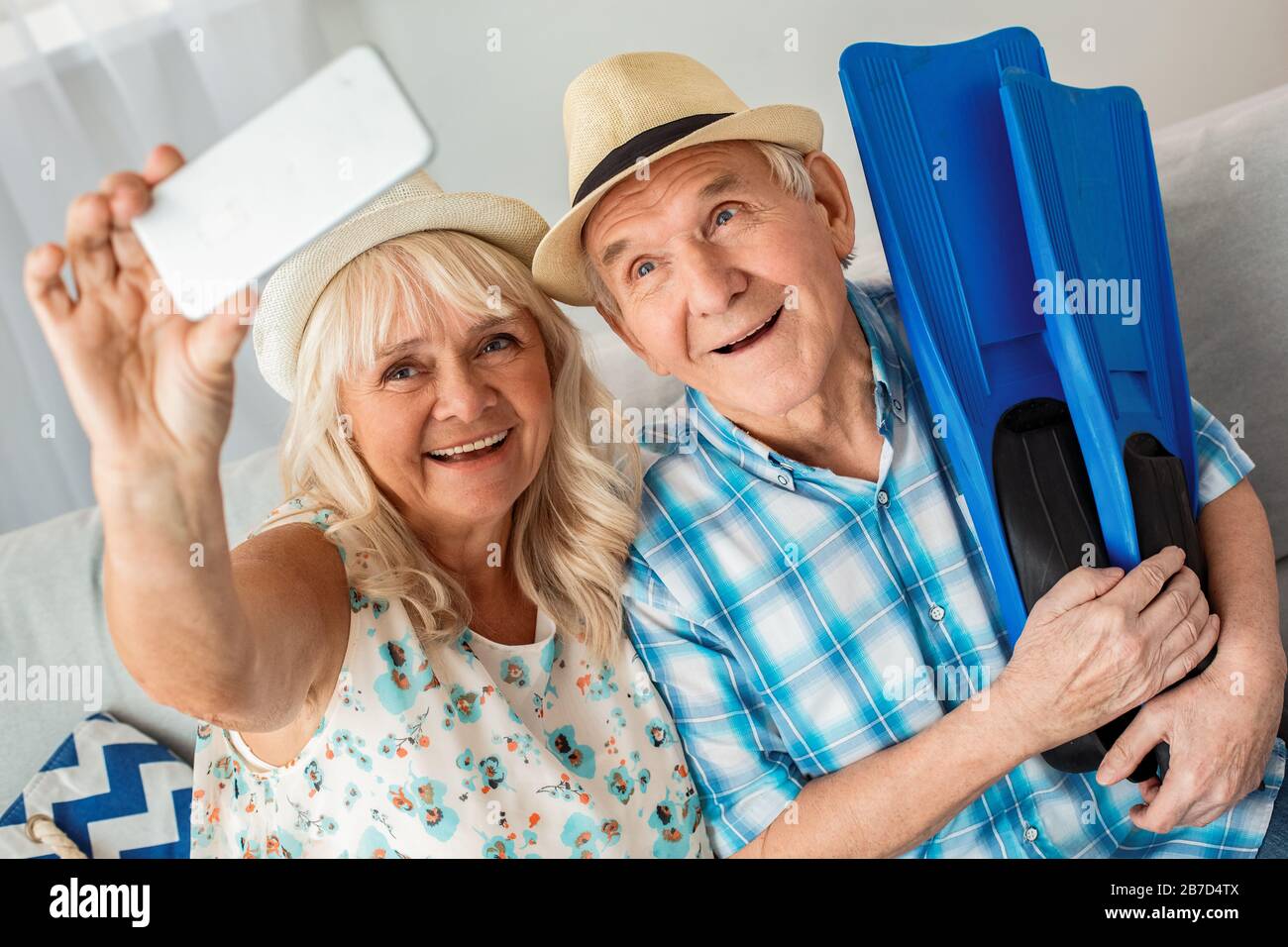 Leitender Mann und Frau sitzen im Reisebüro auf dem Sofa und tragen Strandhüte Ehemann, der Flipper hält, während die Frau das Smartphone bei selfie hält Stockfoto