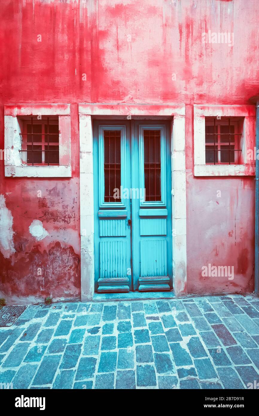 Alte verkratzte blaue Tür und kleine Fenster an bunter roter Hauswand, abstrakte Sicht auf die Stadtstraße mit verlassenen Gebäuden. Schäbige Textur und Grunge Stockfoto
