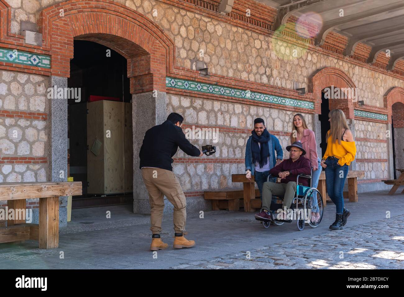 Professioneller Fotograf, der einen jungen Latino mit zwei kaukasischen Mädchen aufzeichnet, die einen paraplegischen erwachsenen Mann im Rollstuhl begleiten Stockfoto