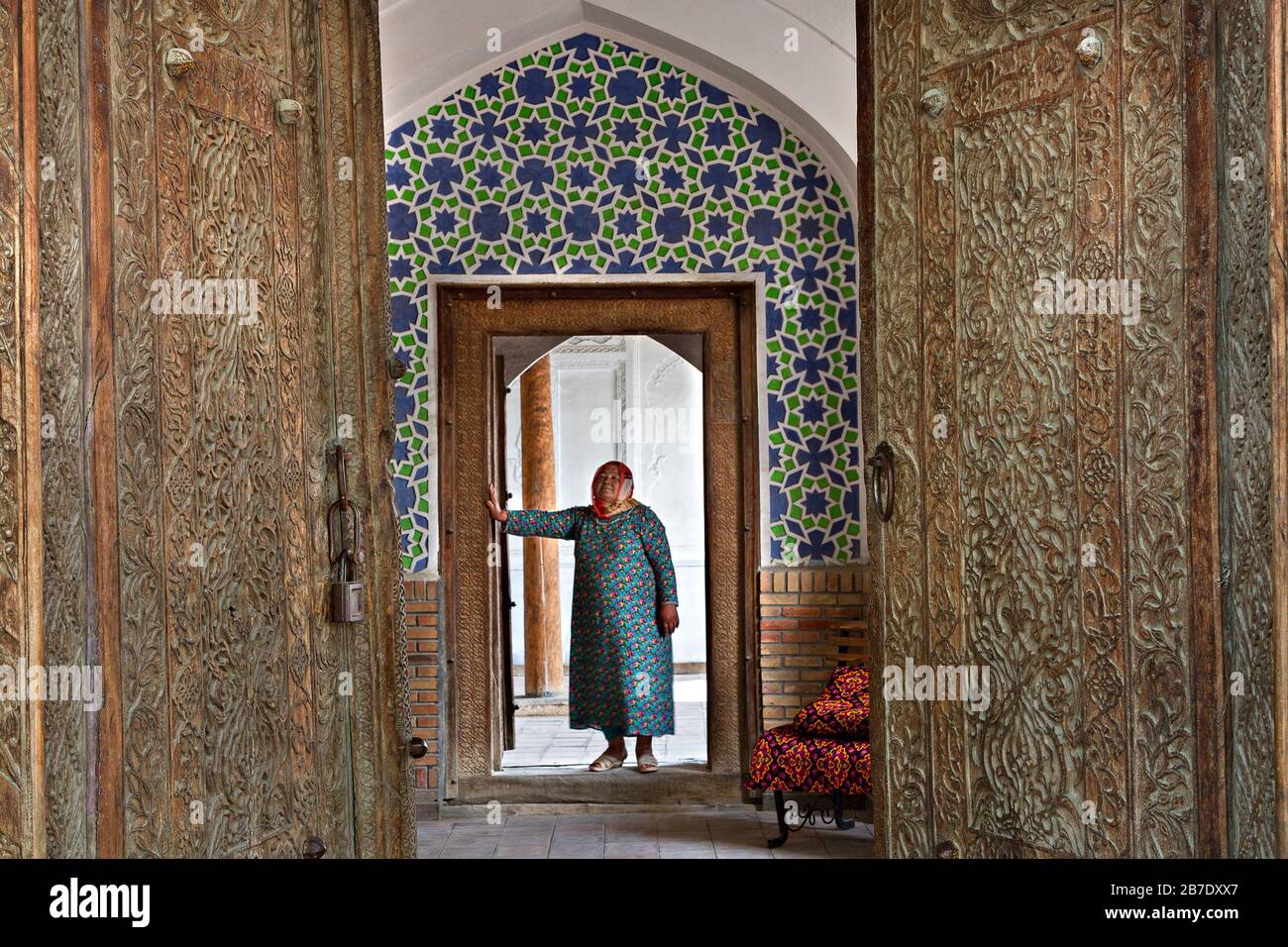 Usbekische Frau in lokaler Kleidung, die durch die Holztüren des Madari Kahn Mausoleums in Kokand, Usbekistan, schaut. Stockfoto