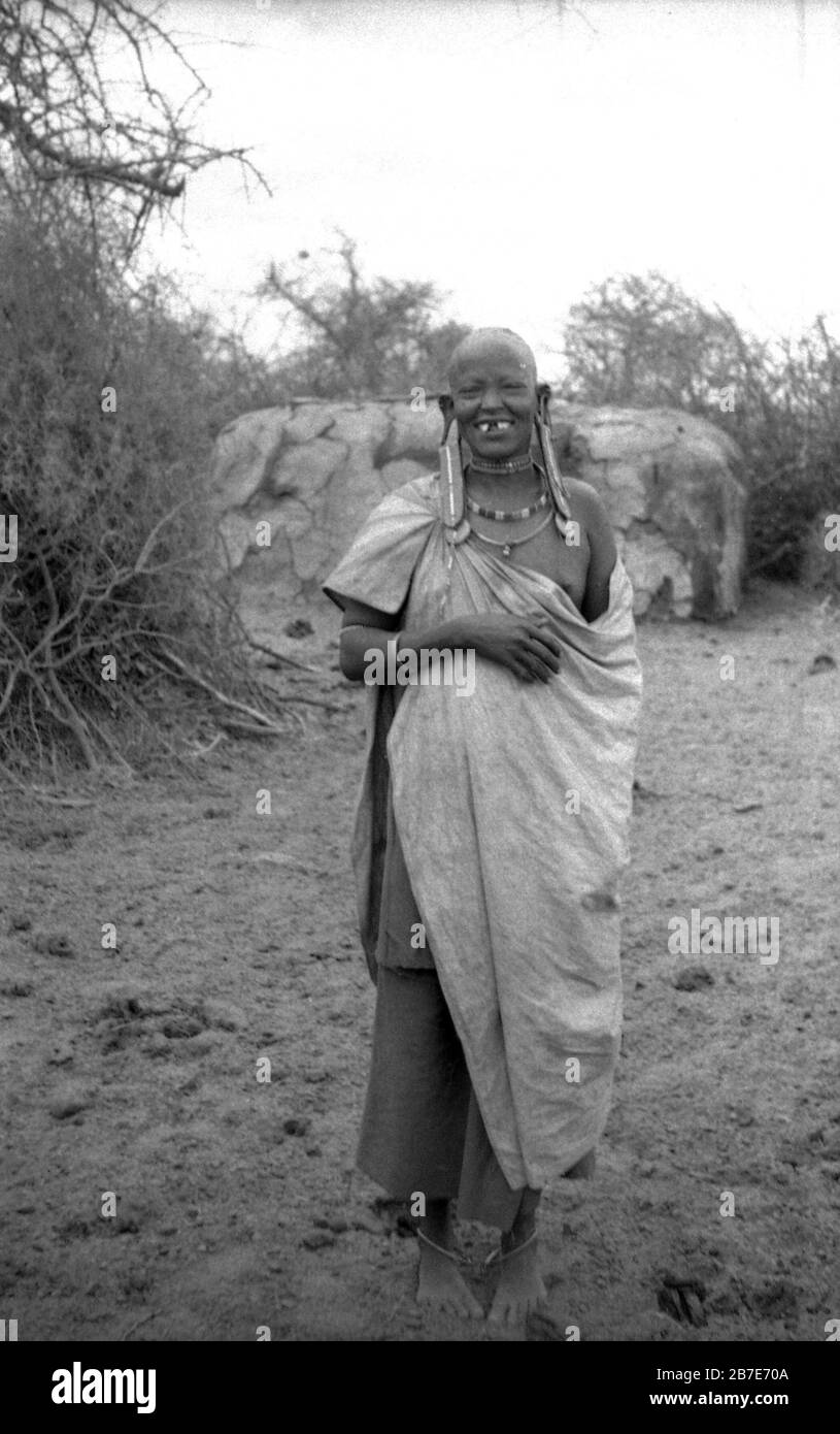 1960er Jahre, historisch, im afrikanischen Busch, eine Maasai-Frau, die lächelnd im traditionellen Kleid steht. Die Masssai sind eine nilotische Gruppe von Menschen, die einen halbnomadischen Reiher-Lebensstil leben und die Maa langage sprechen. Sie bewohnen die afrikanische Großseeregion. Stockfoto