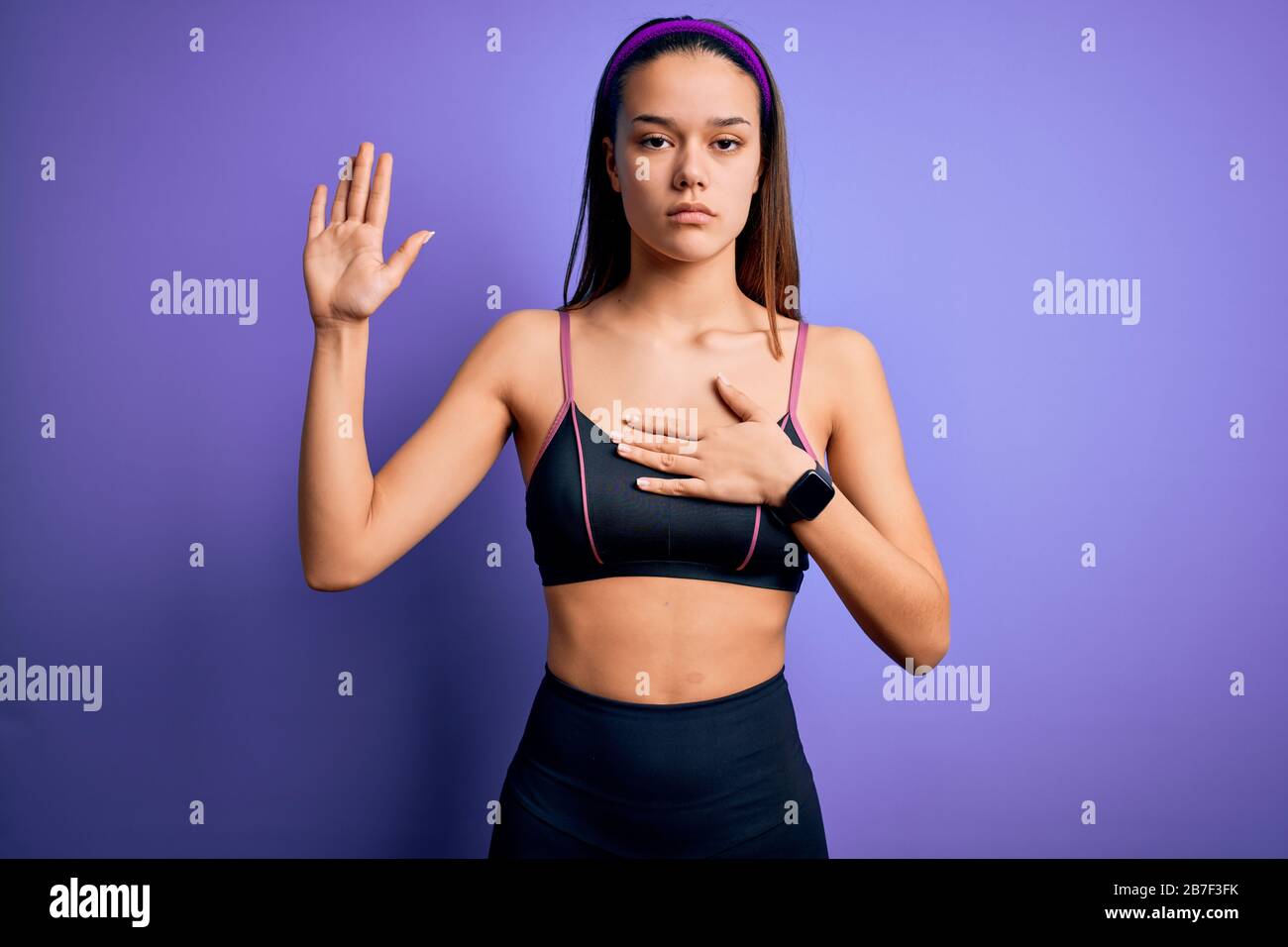 Junge, schöne sportliche Mädchen, die Sport treiben und Sportswear über dem isolierten violetten Hintergrund Tragen, Die Mit der Hand auf der Brust und der offenen Handfläche Schwänzen und so eine Treue machen Stockfoto