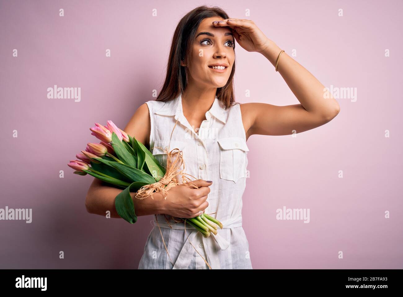 Junge schöne Brunette-Frau, die Blumenstrauß mit Tulpen hält, Blumen über rosa Hintergrund sehr glücklich und lächelnd mit der Hand über Kopf weit weg schauend. Meer Stockfoto