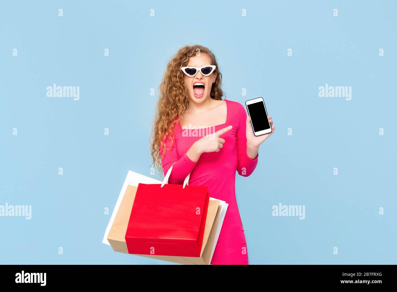 Schockierte Frau mit bunten Einkaufstaschen, die schreien und auf Handy zeigen, isoliert auf blauem Hintergrund Stockfoto