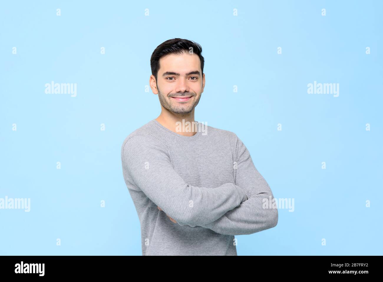 Porträt eines lächelnden freundlichen gutaussehenden Mannes in einfarbig grauem T-Shirt mit Armen, die isoliert in hellblauem Studiobakground gekreuzt sind Stockfoto