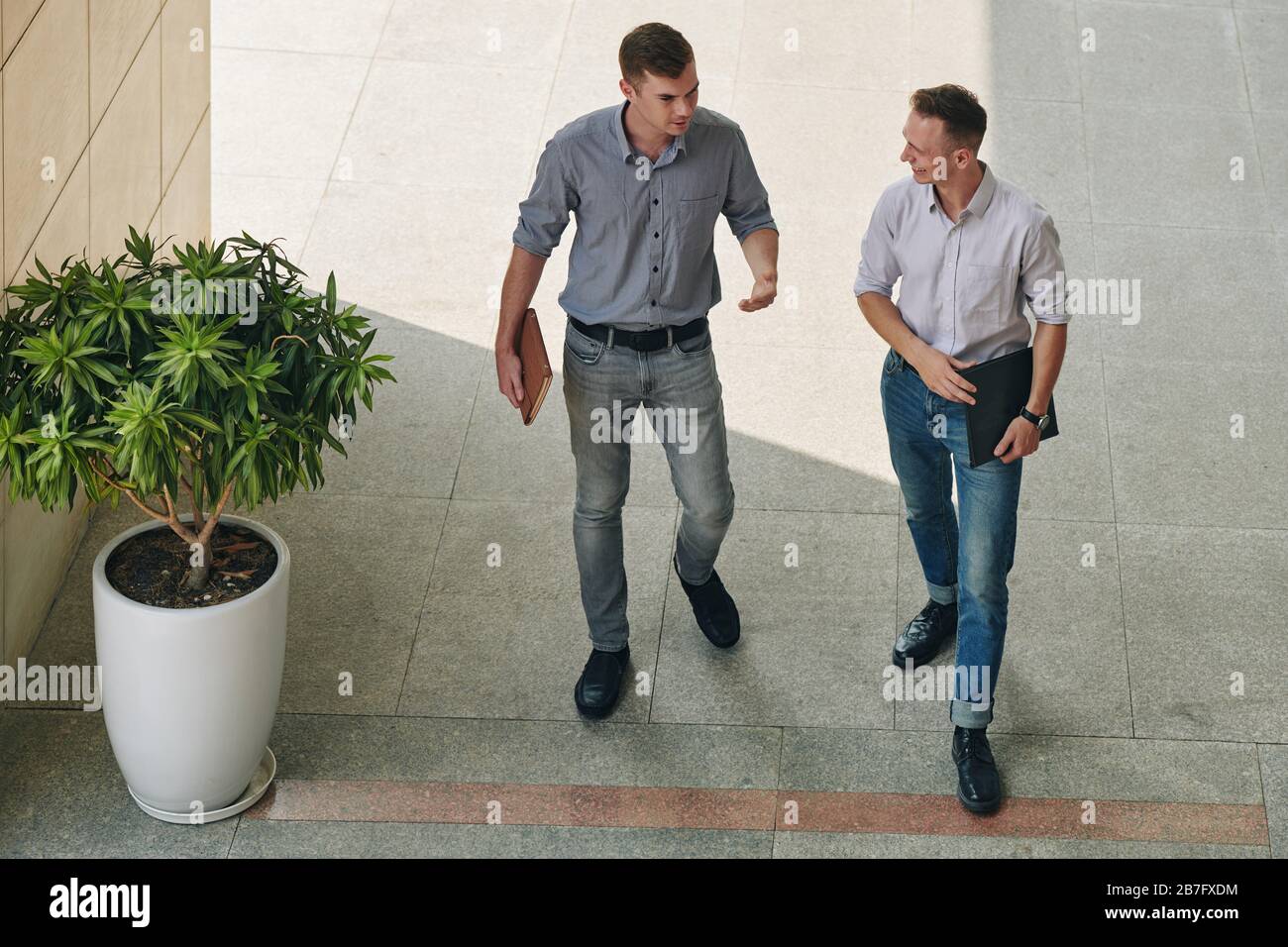 Fröhliche junge Geschäftsleute, die vor dem Bürogebäude spazieren und Nachrichten diskutieren Stockfoto