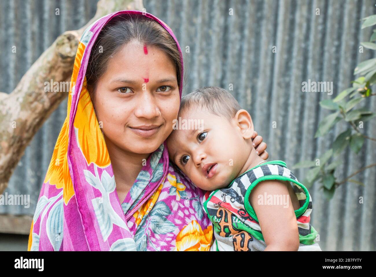 Eine lächelnde Mutter mit Zinnoberrot auf der Stirn steht mit ihrem Baby vor ihrem Haus im Bezirk Bagerhat im Süden von Bangladesch. Stockfoto
