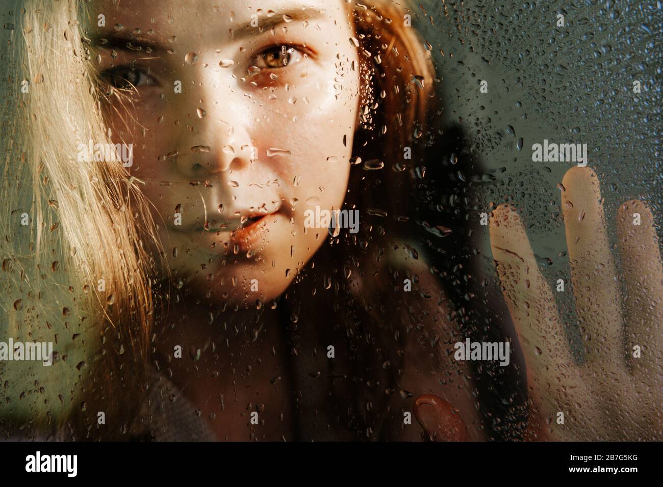 Kaplin von Wasser auf Glas im Fokus. Hinter dem Glas eine verschwommene Silhouette eines Mädchens. Das Mädchen schaut in den Rahmen. Stockfoto