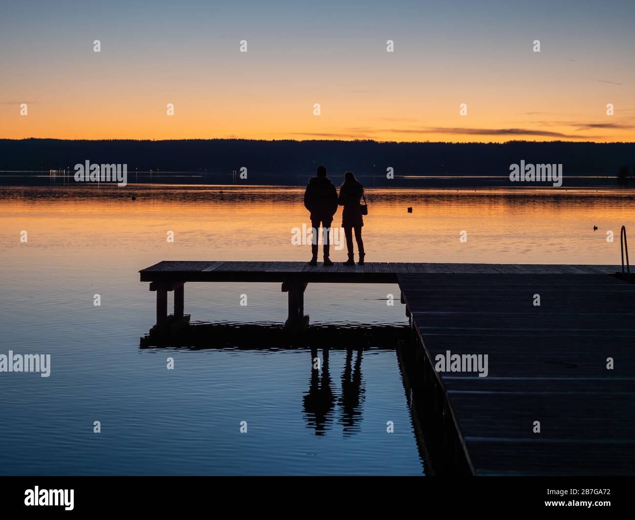 Rückansicht eines Paares, das auf Holzsteg am Bavaria Ammer bei Sonnenuntergang mit alpen im Hintergrund steht Stockfoto