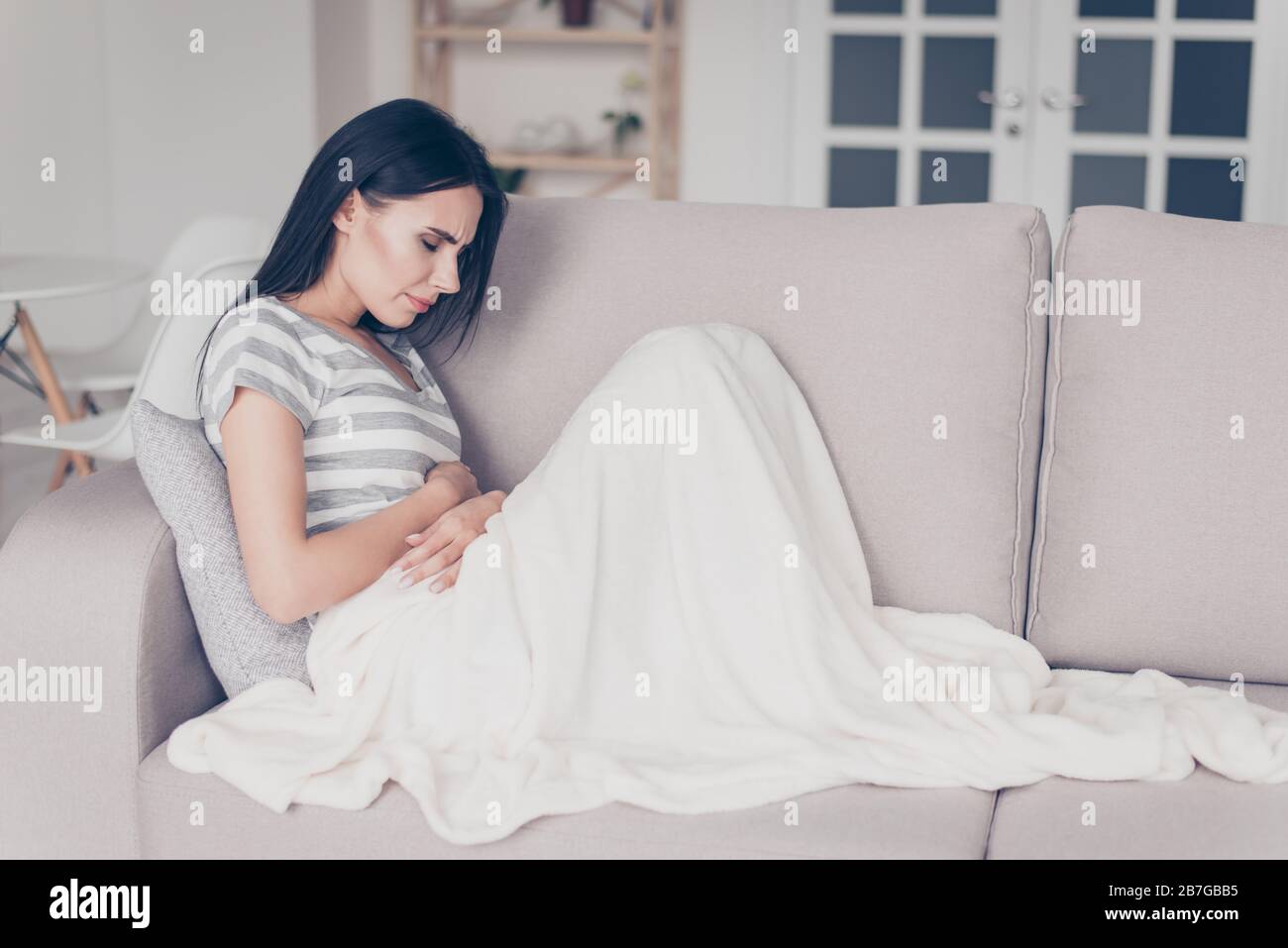 Traurige, schöne Frau, die auf der Couch sitzt und Spasmus und Symptome von pms spüren, während sie mit Decklack den Bauch berührt Stockfoto