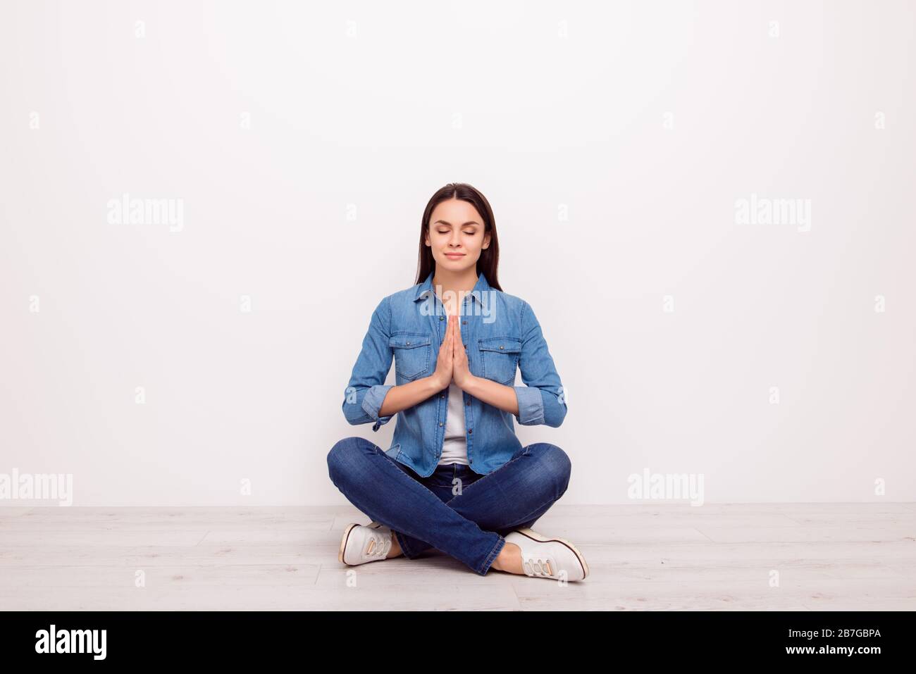 Ruhige, entspannte, hübsche Mädchen in legeren Jeansbekleidung, die Hände hält, geschlossene Augen, in Yoga-Pose sitzend mit gekreuzten Beinen auf dem weißen Boden Stockfoto