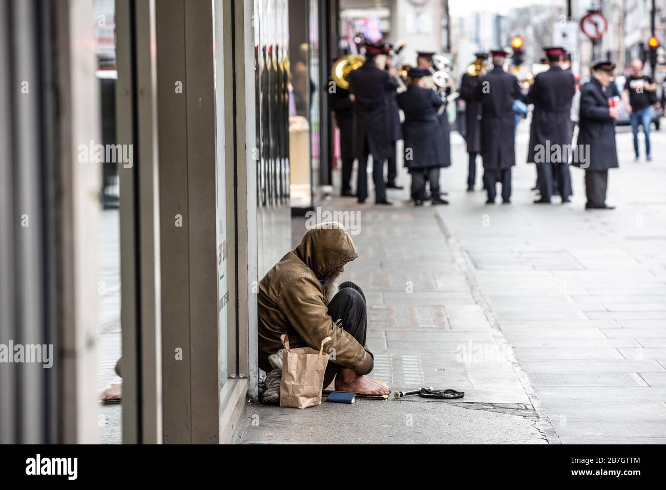 Obdachloser sitzt an einem Schaufenster, während die Heilsarmee Band in der Ferne Passanten in der Oxford Street, London, UK, Musik spielt Stockfoto