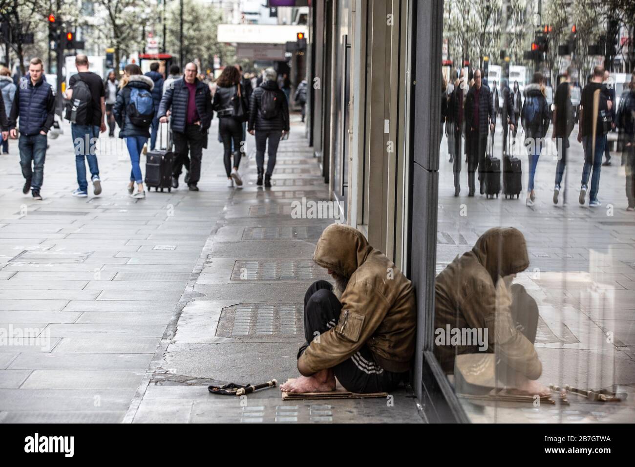 Obdachloser sitzt an einem Schaufenster, während die Heilsarmee Band in der Ferne Passanten in der Oxford Street, London, UK, Musik spielt Stockfoto