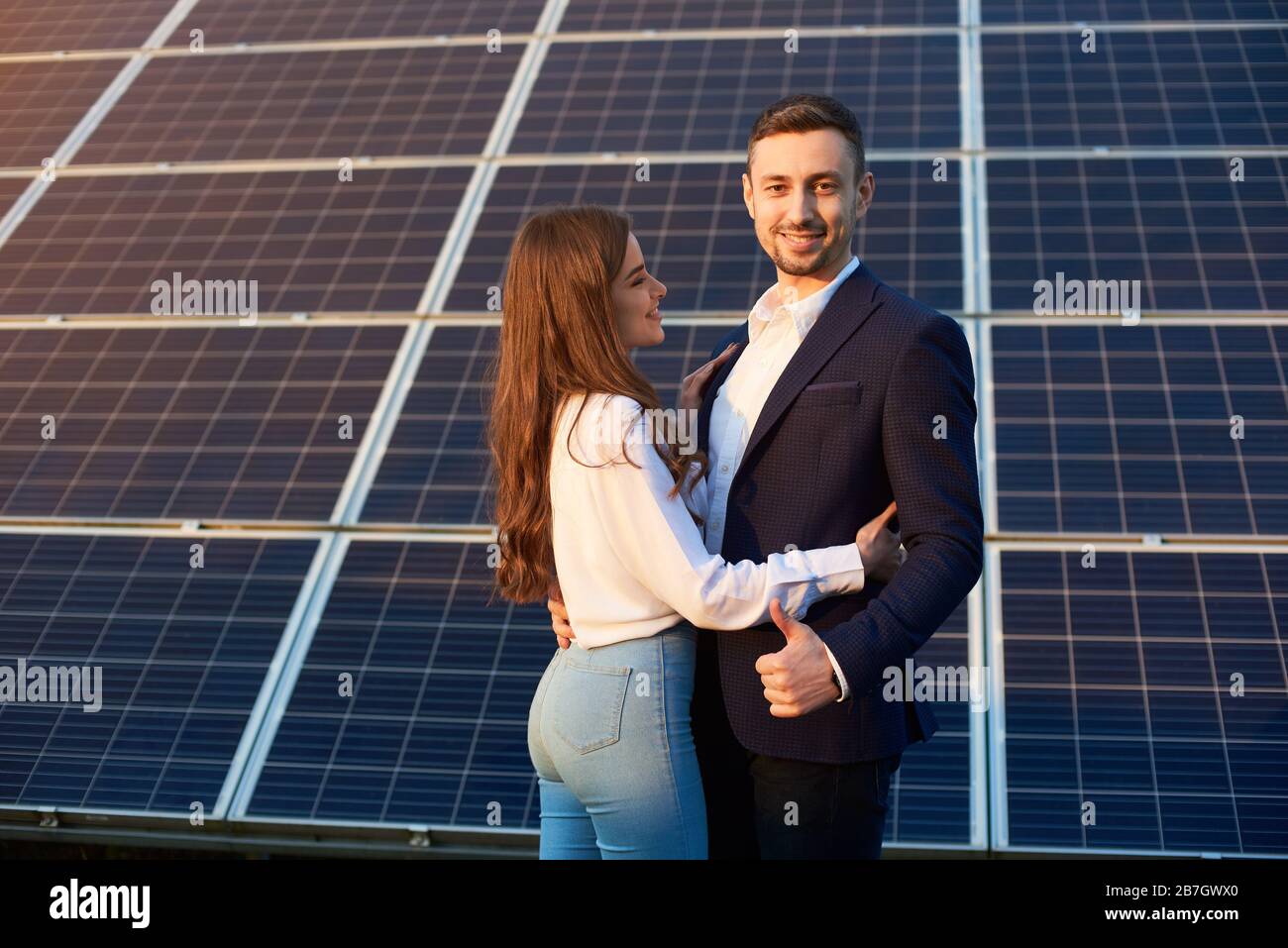 Wunderschönes Paar, das sich auf dem Hintergrund von Solarpaneelen umarmt. Schlankes Mädchen in Jeans mit langem Haar sieht mit Bewunderung auf einen Kerl in einer Jacke aus, der wie ein Daumenschild zeigt. Bild des Solarenergiekonzepts Stockfoto