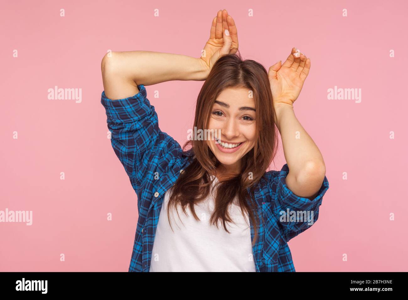 Ich bin Kaninchen! Porträt eines verspielten, hübschen Mädchens in kariertem Hemd, das mit Hunny Ears Geste zeigt, lacht und lustige, kindliche unbeschwerte Stimmung hat. i Stockfoto