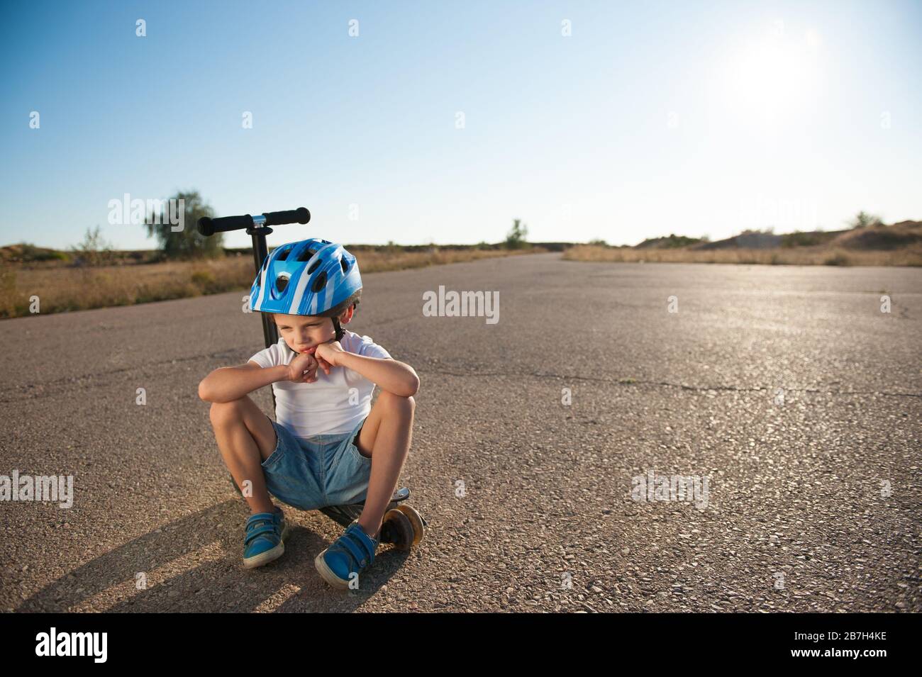 Trauriges kleines aktives Kind im Sport Schutzhelm einsam auf Roller auf leerer Wüstensommerasphalt-Straße bei heißem Sonnenuntergang sitzen Stockfoto