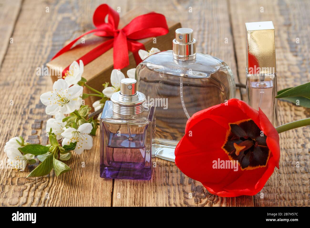 Geschenkschachtel mit rotem Band und Parfümflaschen auf Holzbrettern mit roten Tulpen- und Jasminblüten auf alten Holzbrettern. Stockfoto