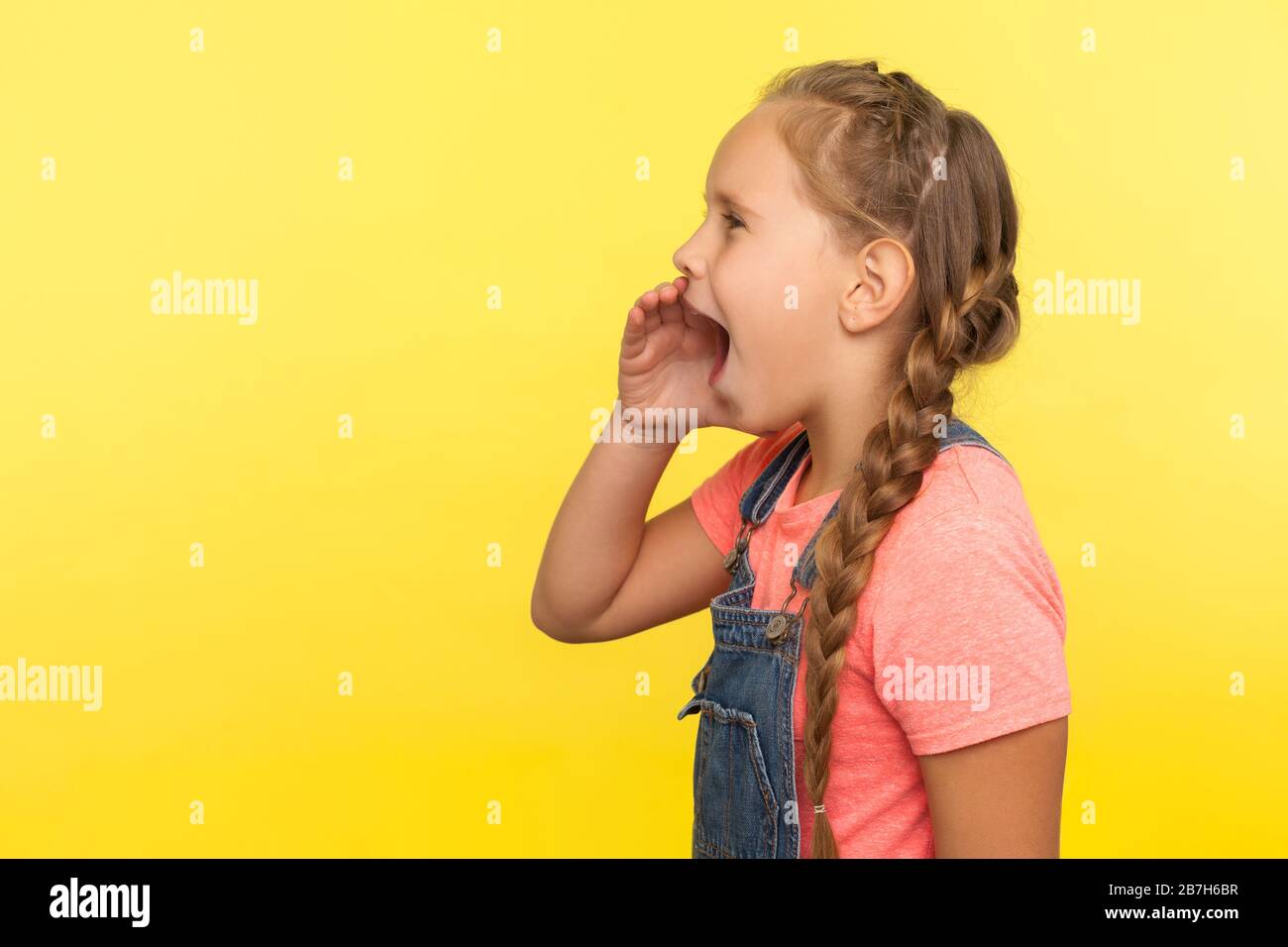 Achtung! Seitenansicht des niedlichen kleinen Mädchens mit Geflecht in Denim-Overalls, die Hand in der Nähe des offenen Mundes halten und laut schreien, Ankündigung schreien Stockfoto