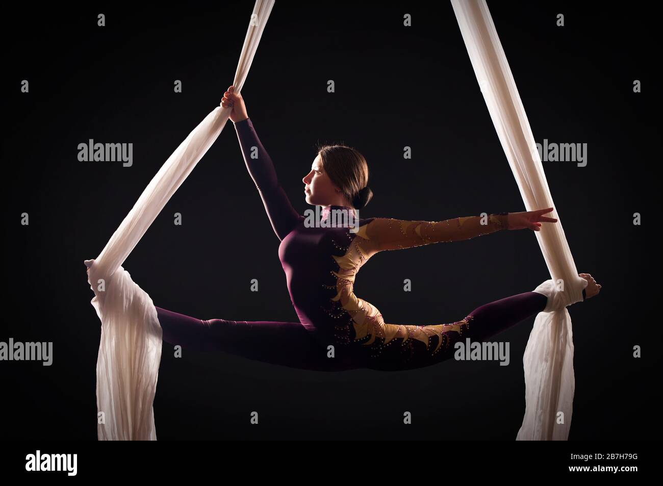 Sportliche Frau im Burgund Anzug führt Gymnastik- und Zirkusübungen auf weißer Seide im Kontralicht durch. Studioaufnahmen vor dunklem Hintergrund. Stockfoto