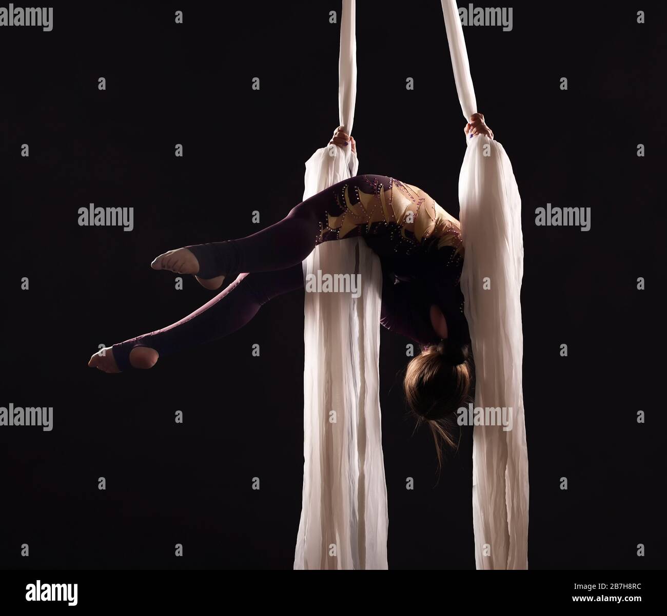 Sportliche Frau im Burgund Anzug führt Gymnastik- und Zirkusübungen auf weißer Seide im Kontralicht durch. Studioaufnahmen vor dunklem Hintergrund. Stockfoto