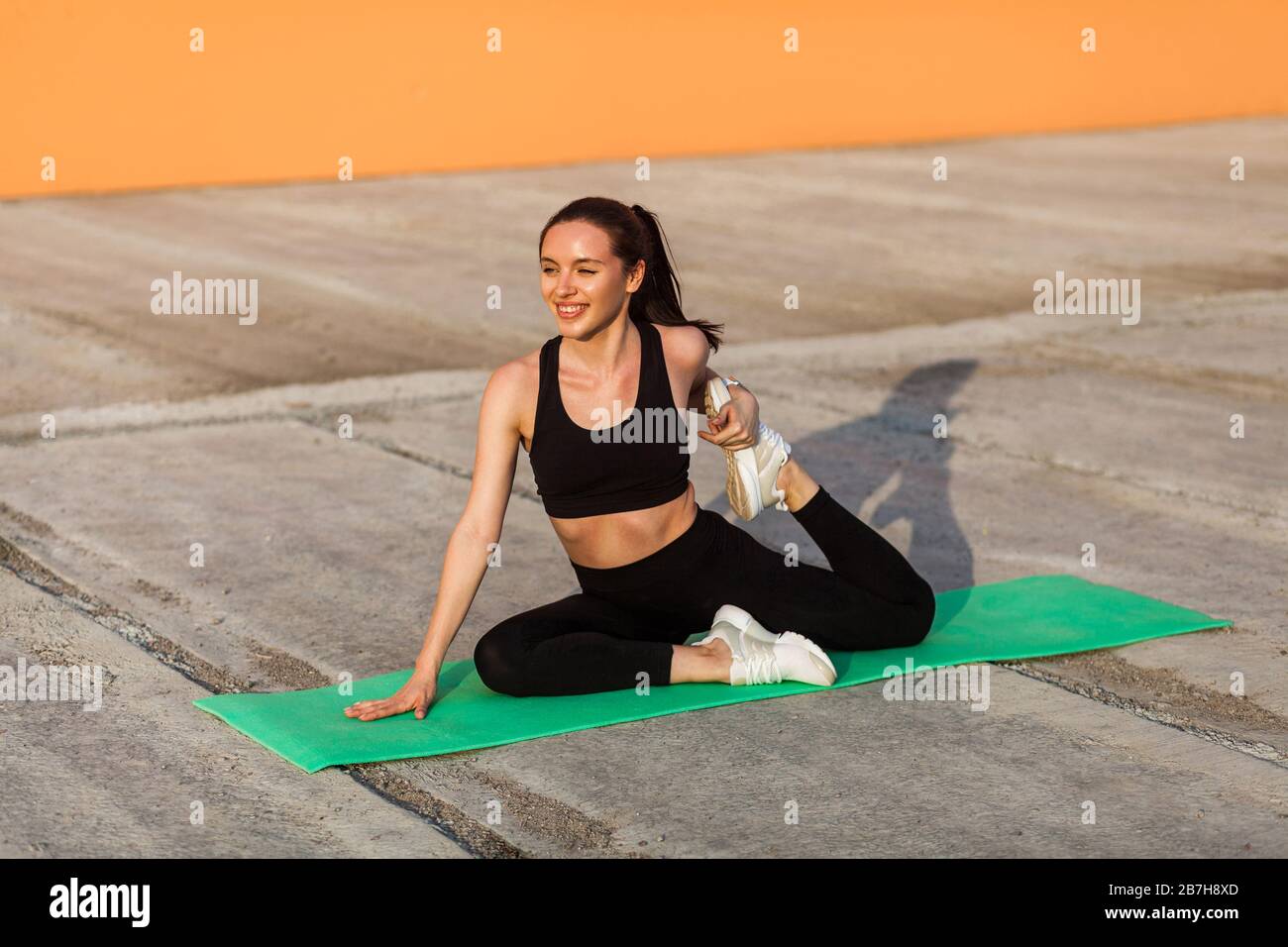 Lächelndes sportliches Mädchen in enger Sportswear, schwarzer Hose und Oberteil, praktizierendes Yoga, einbeinige Königstaubenpose machen und Beinmuskeln für bessere Strecken Stockfoto